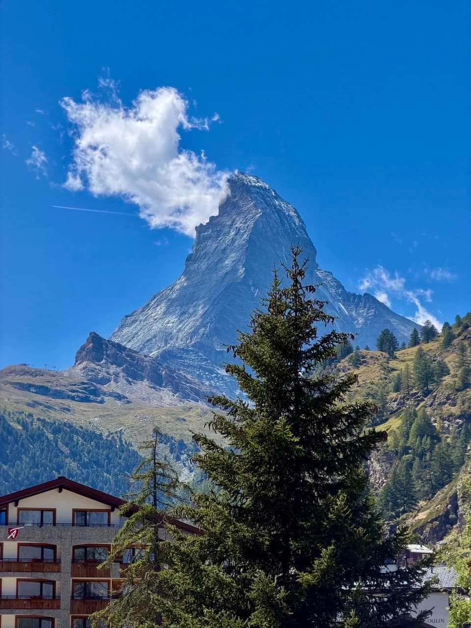 A white cloud curling around the Matterhorn beneath a blue sky, with Alpine slopes and a fir tree below