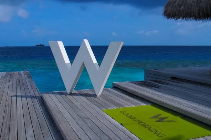 A white W sculpture and lime-green Good Morning mat on a timber deck beside the turquoise sea at W Maldives