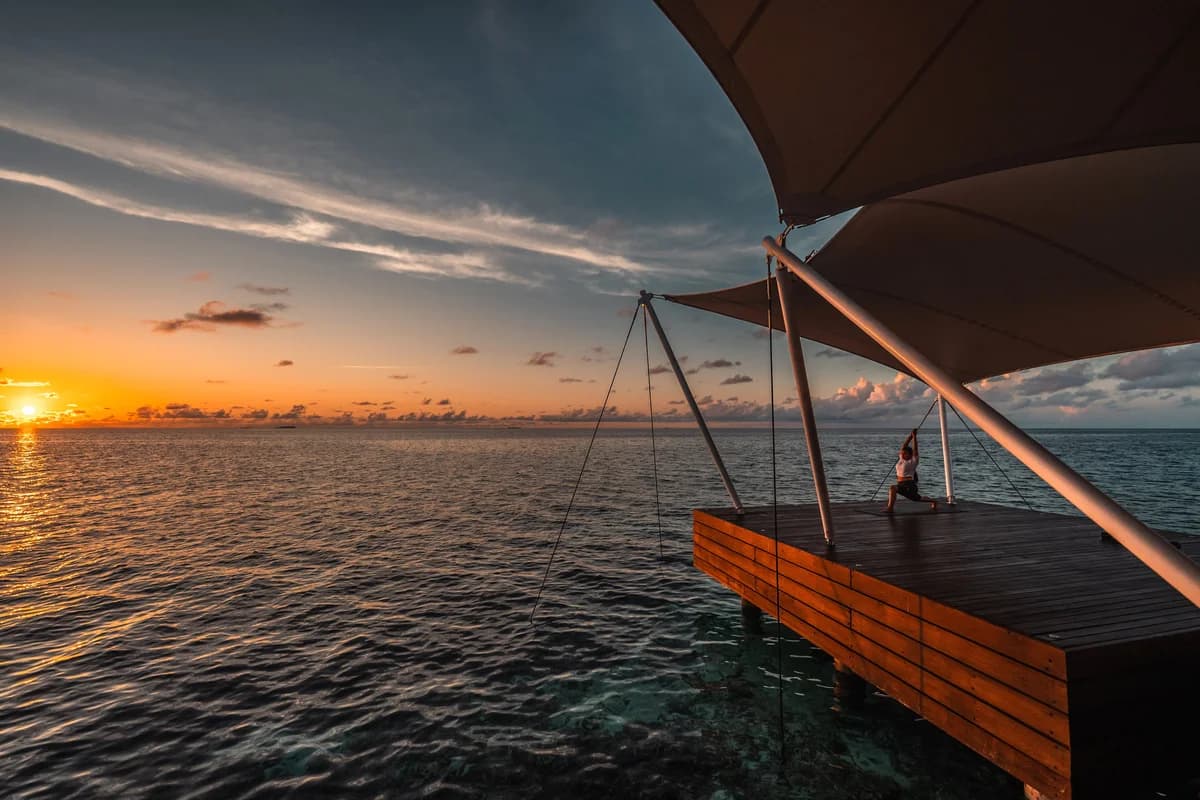 Yoga beneath a sail canopy on an overwater deck at W Maldives, with the sun setting over the ocean
